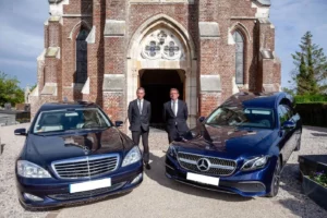 Deux agents funèbres en costume posant avec deux corbillards Mercedes bleus devant l'église