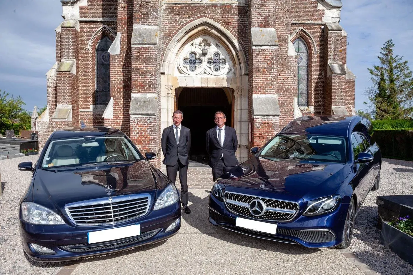 Deux agents funèbres en costume posant avec deux corbillards Mercedes bleus devant l'église